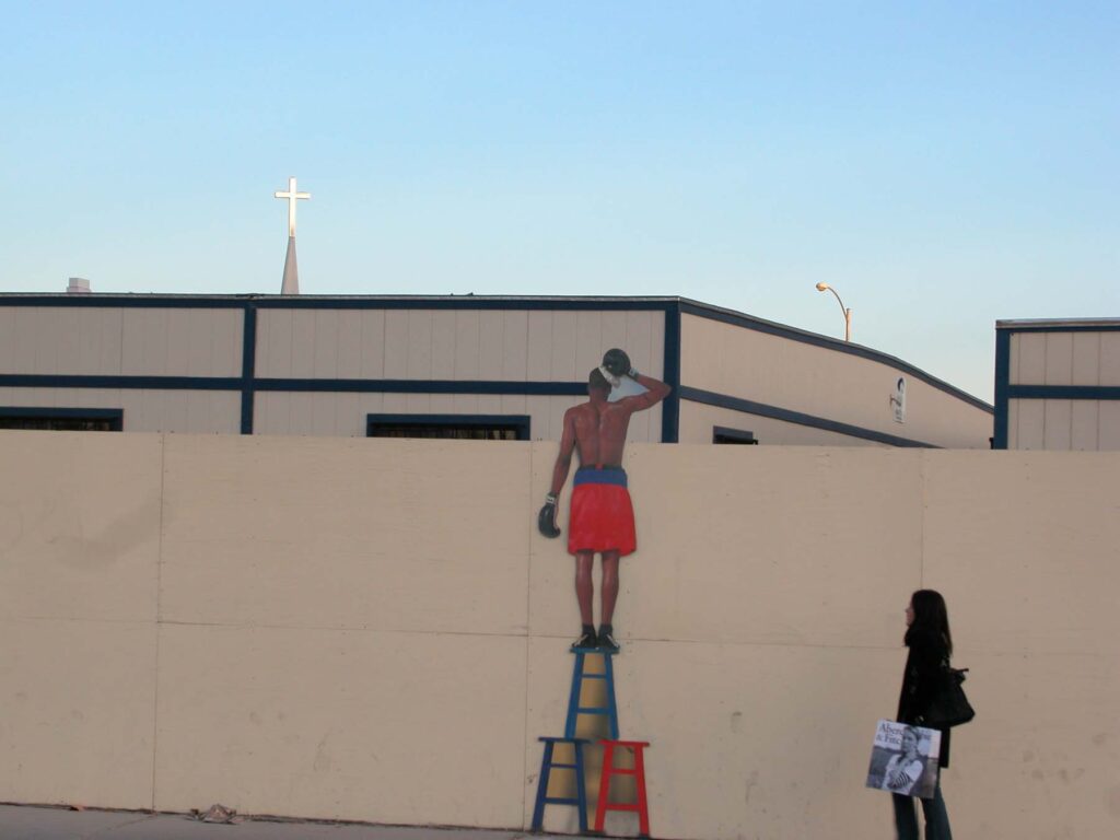 Mural of man on ladder looking up toward a cross jutting above nearby buildings.