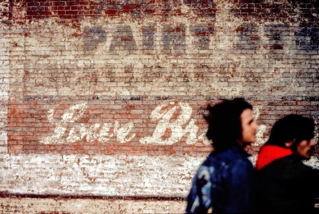 Two motorcycle riders in front of an old brick storefront with words Lowe Brothers, photographed in Davenport, Iowa, 1977.